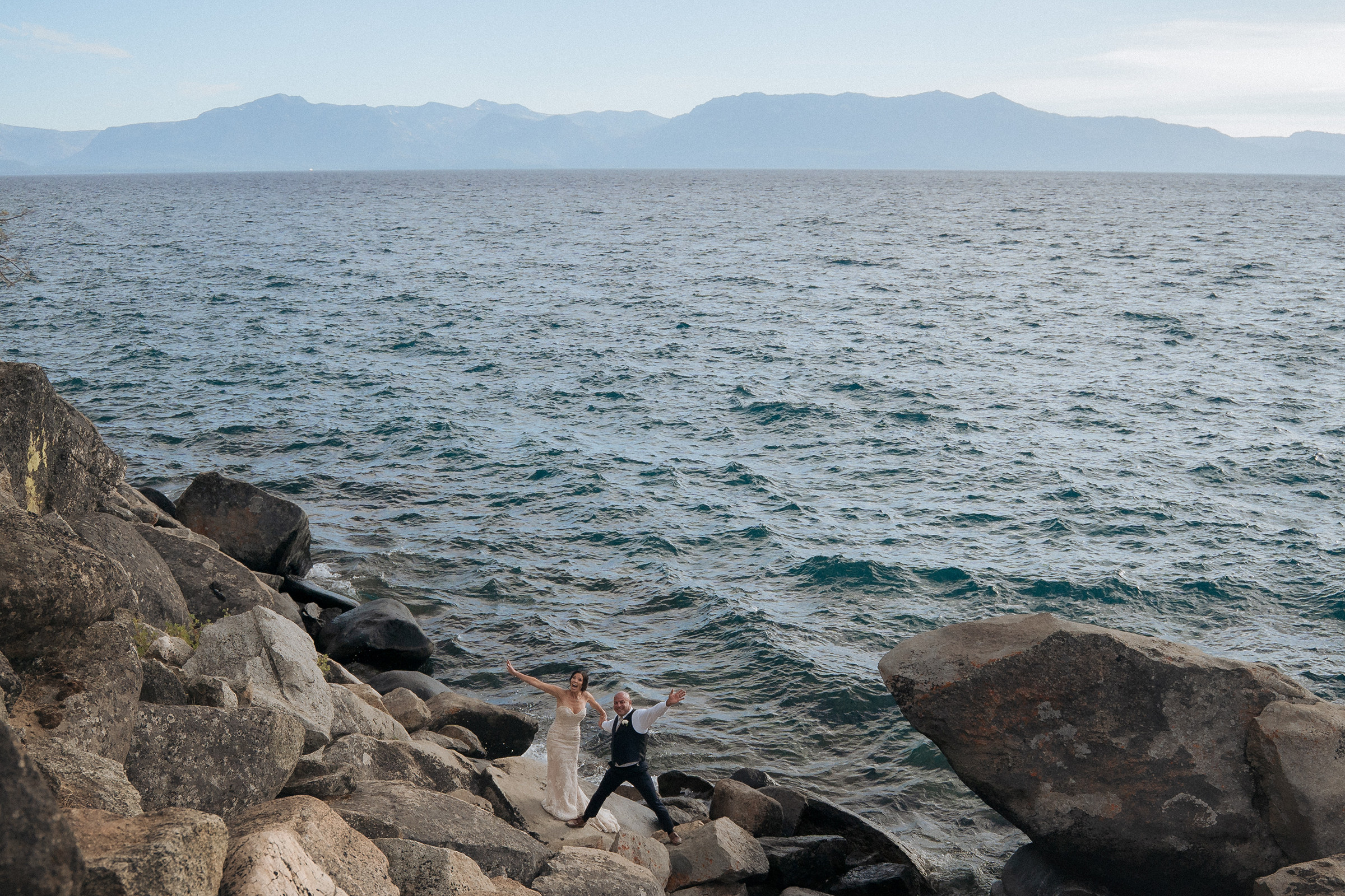 Logan Shoals elopement couple standing along the rocky Lake Tahoe shoreline with expansive water and mountain views in the background.