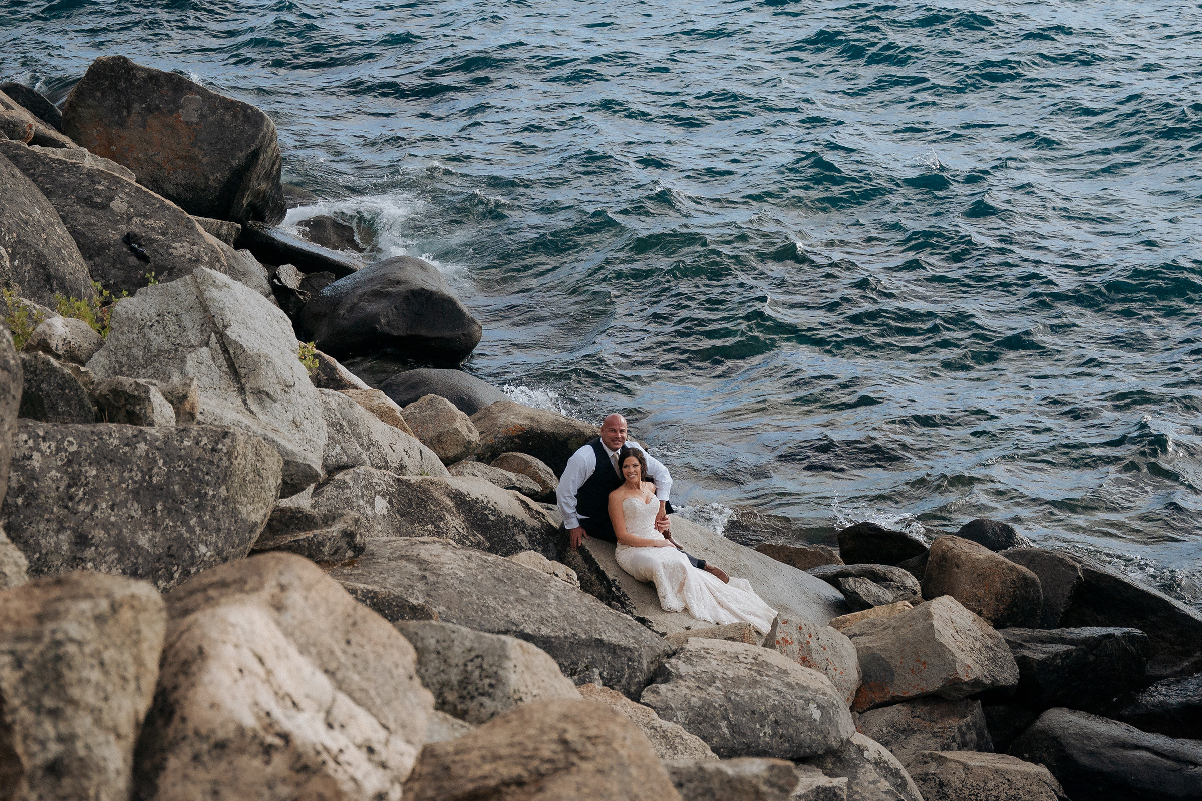 Logan Shoals Lake Tahoe elopement couple embracing along the rocky shoreline with scenic lake views and mountains in the distance.